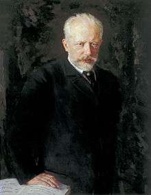 A middle-aged man with grey hair and a beard, wearing a dark suit and staring intently at the viewer