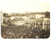 An old photograph showing a crowded square in front of a large, white, multi-storied building