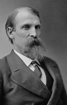 Black and white photograph of a serious-looking middle-age man with thinning hair, long mustache, and pointy beard, wearing a double-breasted waistcoat and necktie