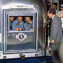 Photo of the three crew members smiling at the President through the glass window of their quarantine chamber. President Nixon is standing at a microphone, also smiling.