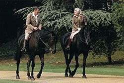 Elizabeth and Ronald Reagan on black horses. He bare-headed; she in a headscarf; both in tweeds, jodhpurs and riding boots.