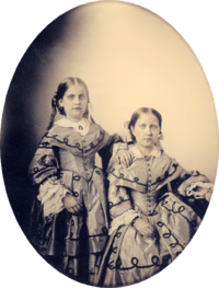 An oval, framed photographic portrait of two young girls dressed in elaborate Victorian-era gowns