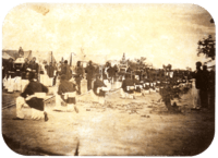 An old photograph showing a procession passing between lines of soldiers with tents in the background