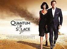 A man in a business suit holding a gun with a woman in a black dress walking away from a destroyed facility in the desert. To their left is the title "Quantum Of Solace" in black letters – except the 'O's, which are golden and make a diagonal straight line with a 7 resembling a gun.