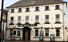 Front of a three-storey building, six windows across, with a large-framed wood door at ground level and a painted sign with the words "THE QUEENS HEAD"