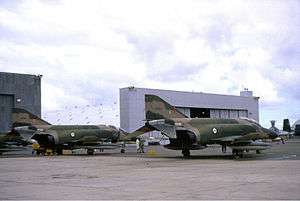Colour photo of two military jet fighters painted in a camouflage pattern parked in front of a large white building