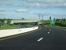 Ground-level view of a divided freeway at a curve in the road; a green exit sign and a white bridge are visible in the distance.