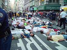 Street protest scene; people deliberately lying down on a busy city street, surrounded by onlookers and police