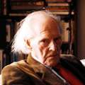 Photographic portrait of an unsmiling, elderly man looking side on into camera. The hair he has is white and collar length. He wears a brown jacket, blue collared shirt and red tie. He appears to be seated and in front of a bookcase.