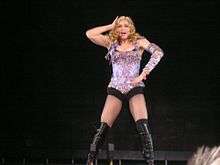 Image of a blonde woman. She is wearing a purple sparkling corset with black shorts and black knee-high boots. She has her right arm to her hip and her left arm to her head.