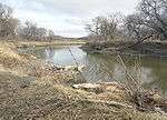 A small river flows through a prairie landscape; brown grasses and leafless trees line the banks.