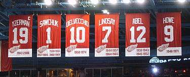 The banners of six retired numbers. The banners, from left to right, read "Yzerman 19" "Sawchuk 1" "Delvecchio 10" "Lindsay 7" "Abel 12" "Howe 9". The Yzerman banner has a small "C" at the top right corner.