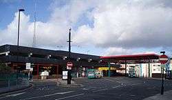 A 1980s multi-storey car park with a large tarmac bus area and light railway station. Two buses can be seen in the station. A red coloured roof is over a section of the bus and metro station and the metro station has its own roof coloured in cadmium yellow.