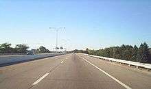 Ground-level view of two lanes of a divided freeway; the opposing lanes of traffic are separated by a narrow concrete barrier. A dense forest is visible to the left.