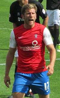 A man with short brown hair is wearing a red top, blue shorts and white socks. He is standing on a grass field.