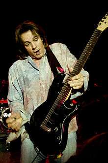 A fifty-five-year-old man is holding an electric guitar in his left hand more than mid-way down its neck. His right hand has a plectrum between thumb and forefinger, it is poised above and to the left of the guitar. The man has dark hair and is open mouthed while looking down at the guitar. Behind him is an obscured drum stand and other band equipment.