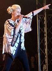 Young blonde woman singing onstage, pointing upwards