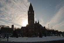 A fine and ornate building with a huge clock tower fills the central part of the image. It appears directly in front of the sun giving it a dark silhouette-like setting. The scene is marked by white snow.