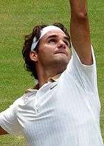 A brown-haired man in a white polo shirt raises his left arm, preparing to serve