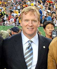 A blond man with a dark blue suit, a white shirt, and a striped blue tie, standing before a crowd