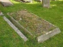 A horizontal gravestone, badly dilapidated and with grass growing among the cracks