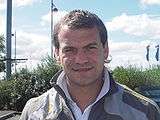 A head-and-shoulders photograph of a man with short brown hair wearing a grey jacket over a white shirt.