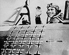 A fighter pilot sitting in an aircraft cockpit, shown in profile, viewed from the left. The pilot is smiling and waving his right hand in the air. The left side of the cockpit bears approximately 25 small black crosses arranged in five rows and five columns.