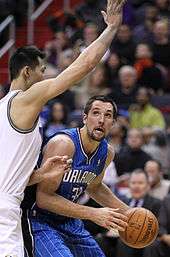 A basketball player, wearing a blue jersey with the word "ORLANDO" and the number 33 on the front, with the basketball and being guarded by a player who has his right arm extended.