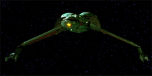 Forward shot of a starship in the black of space with distant stars in the background. The ship is primarily one color, save for a decorative pattern on its underside. The center portion of the ship is bulbous, with thin, down-stretched wings capped by long guns.