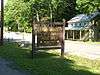 A brown wooden sign with yellow letters reading "Sand Bridge State Park Picnic Area Pennsylvania Department of Conservation and Natural Resources" in front of a highway and pale green two story frame house