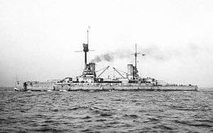 A large warship steaming in choppy water; black smoke drifts up from the ship's two tall funnels