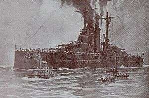 A large battleship sits motionless in the water with smoke coming out of its funnels and three small boats moving beside her in the foreground.