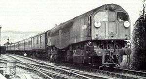 A side-and-front view of an 0-6-6-0T steam locomotive travelling towards the cameraman. The locomotive is a cuboid that contains boiler, fuel and cabs. The cuboid sits on two 0-6-0 bogies. It is hauling a train of around seven carriages past a signal box in the background.