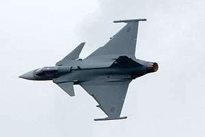 Grey fighter aircraft banking right, showing its top view, against a light-blue sky