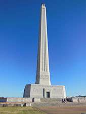 A tall stone column, which widens as it meets the base, a large rectangular building with no windows.