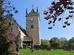 A bright spring day on a country churchyard, surrounded by mature trees.  The church nave is on the extreme left, and at the far end is tall, square, tower capped by a pointed wooden roof, not quite a spire, and with pinacles on all 4 corners.  A bold white clockface is on the tower.  The church has an ope porch, with pillars of wood.  Dark, old, gravestones are scattered in the green grass.