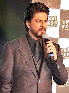 Shah Rukh Khan bearded, in a black suit is interacting with the media