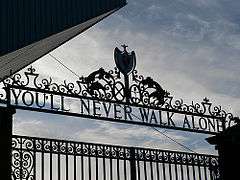 Design of the top of a set of gates, with the sky visible. The inscription on the gates reads "You'll Never Walk Alone".