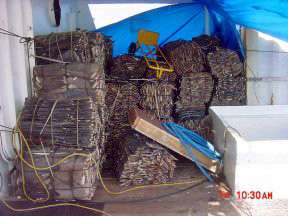 Small tied bales of flat gray objects, messily stacked underneath a translucent tentlike roof between white walls.