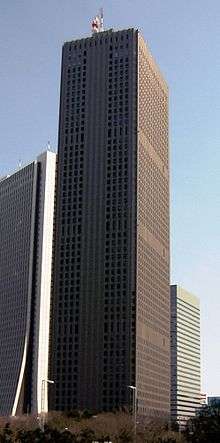 Ground-level view of a brown, rectangular high-rise; the window placement creates several horizontal bands on one side and one vertical stripe on the other