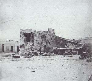 Photograph of a large, ruined masonry tower within a fortress that is pockmarked with large shell holes and two artillery caissons sitting in the foreground