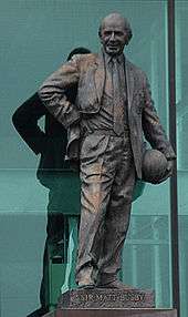 A bronze statue of a smiling bald man holding a football at his left hip and with his right hand on his waist. The plinth reads "Sir Matt Busby".