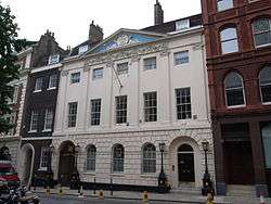 A four storey, white bricked building, with two black doors. A flag pole is protruding from the centre of the building.