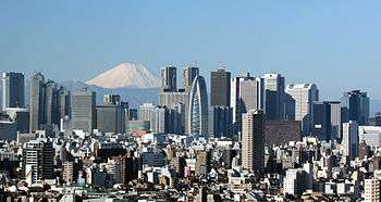 Aerial view of a city; many low-rise buildings in the foreground with many high-rise buildings in the background. The summit of a snow covered mountain can be seen in the distance.