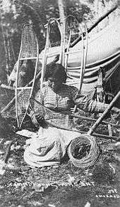 "Black and white photo of a woman kneeling on the ground making a snowshoe. She is wearing a plaid shirt and white dress locking down at the snowshoe. Around her is four frames of snowshoe to be made leaning on a tippy,