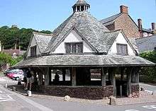 A small single-story building with a pyramid shaped roof, to the side of a road lined with buildings. Some private small cars visible. Trees in the distance with the skyline of Dunster Castle.