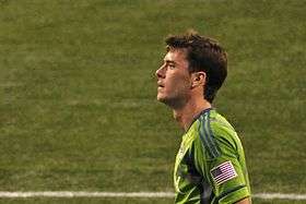 A grown Caucasian man in a light green shirt stares to the camera's left against a green background