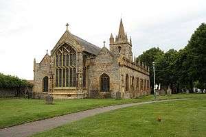 A stone church seen from the north east, with a large Perpendicular east window, aisles with pinnacles, and a west tower with pinnacles and a spire