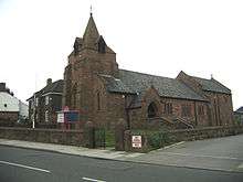 A church seen from the southwest with a low broach spire, the chancel higher than the nave and the porch protruding from the south; in front is a wall, a notice board and a flagpole; to the left a house