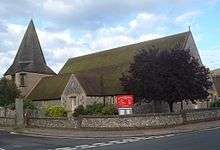 Three-quarter view of a long flint church with an extremely wide, stumpy tower on the left. This is topped with a dumpy grey spire.  The brown church roof is heavily discoloured.  A tree in full leaf obscures the near corner.